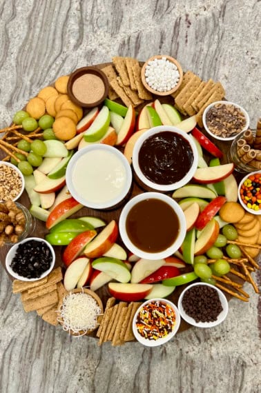 A large round food board filled with slices of red dandelion green apples and an assortment of dipping sauces and garnishes.