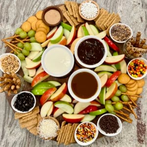 A large round food board filled with slices of red dandelion green apples and an assortment of dipping sauces and garnishes.