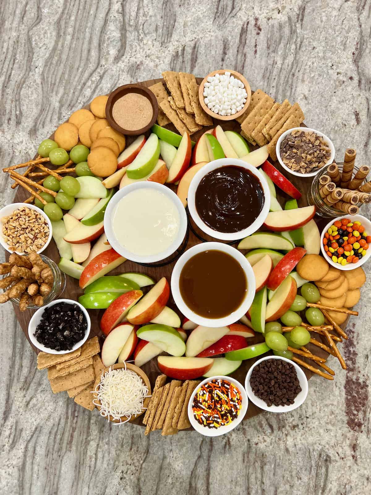 A large round food board filled with slices of red dandelion green apples and an assortment of dipping sauces and garnishes.