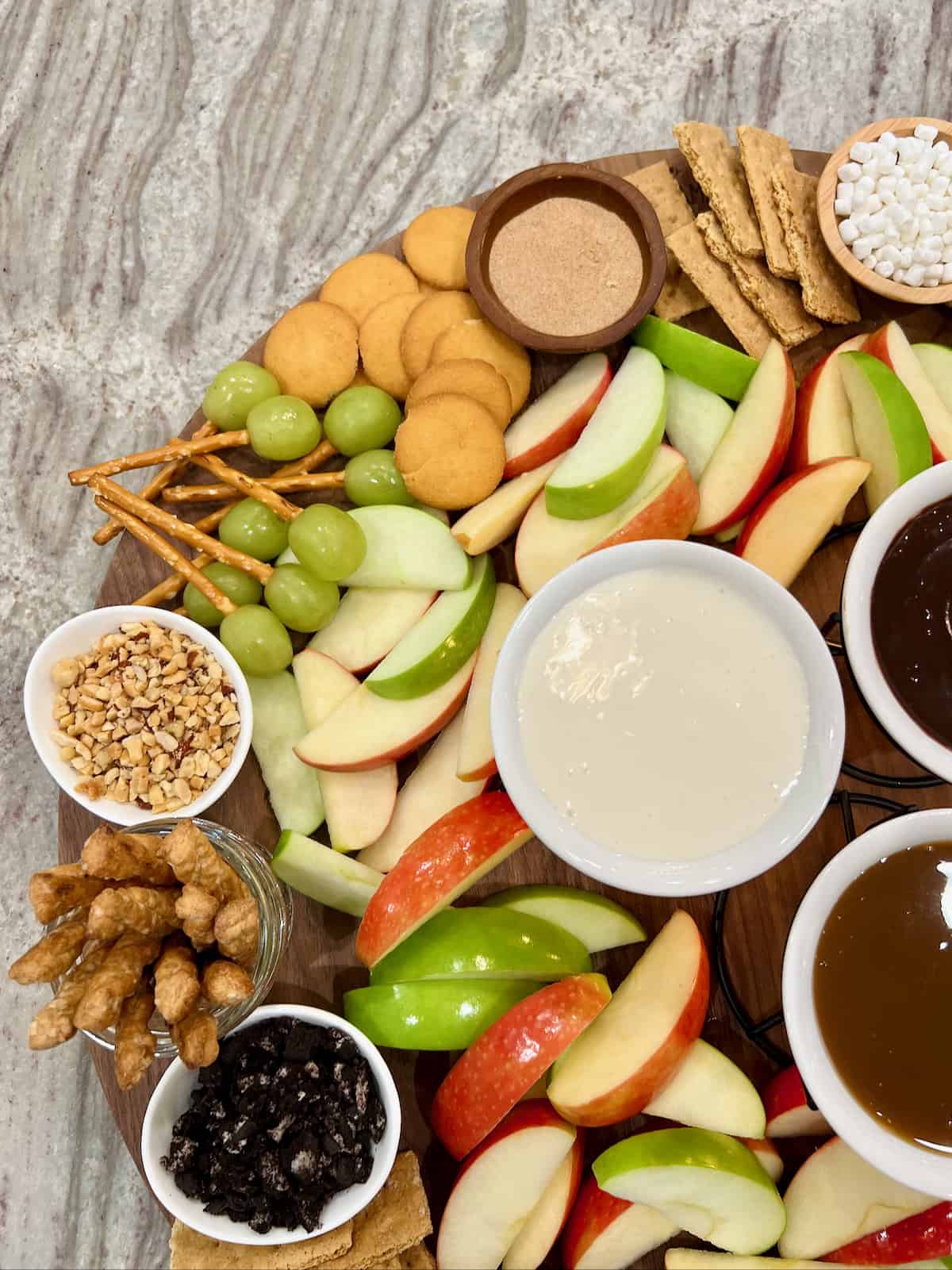 Grapes with pretzels stuck into them next to Nilla wafers and a small white bowl of crushed nuts on a large round wood food board.