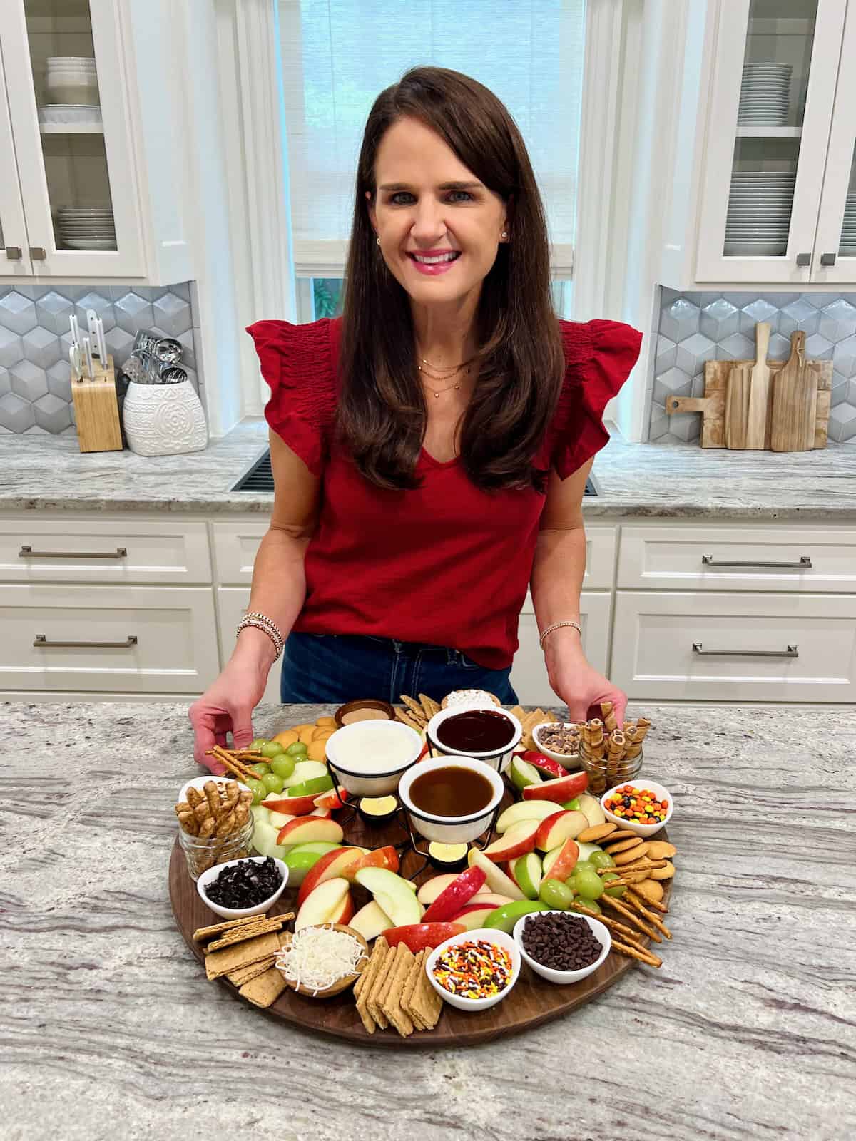 Maegan Brown wearing a red flutter sleeve shirt standing at a gray and white marble counter showing an apple dipping board.