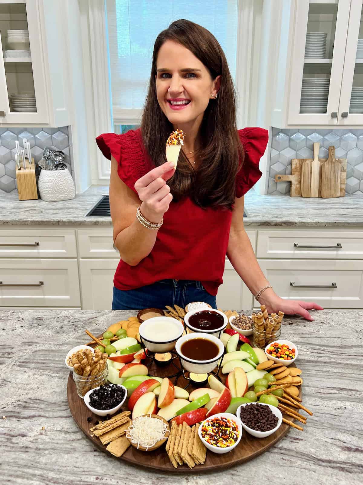 Maegan Brown wearing a red flutter sleeve shirt standing at a gray and white marble counter showing an apple dipping board.