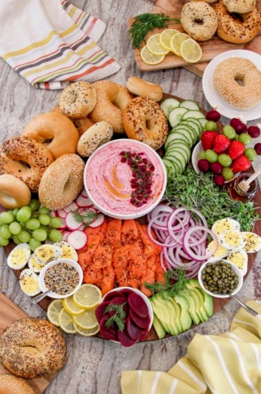 A large round wood board with fixings to make bagel breakfast sandwiches and a white bowl of beet cream cheese in the middle.