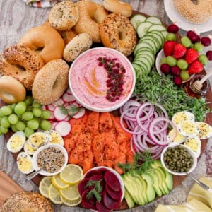 A large round wood board with fixings to make bagel breakfast sandwiches and a white bowl of beet cream cheese in the middle.