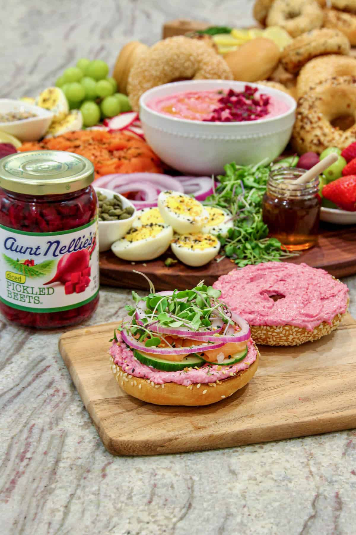 A bagel breakfast sandwich made with beet cream cheese. spread and fresh vegetables and lox on a small board with a jar of pickled beets to the left of it.