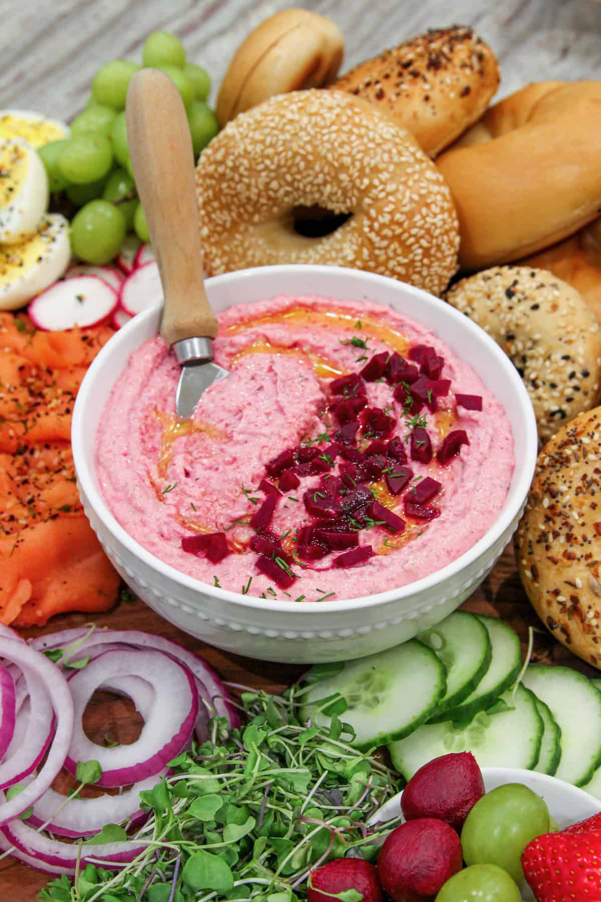 A white bowl of beet cream cheese spread in the middle of a breakfast bagel sandwich board surrounded by fixings to make a bagel sandwich. There is a spread knife in the bowl.