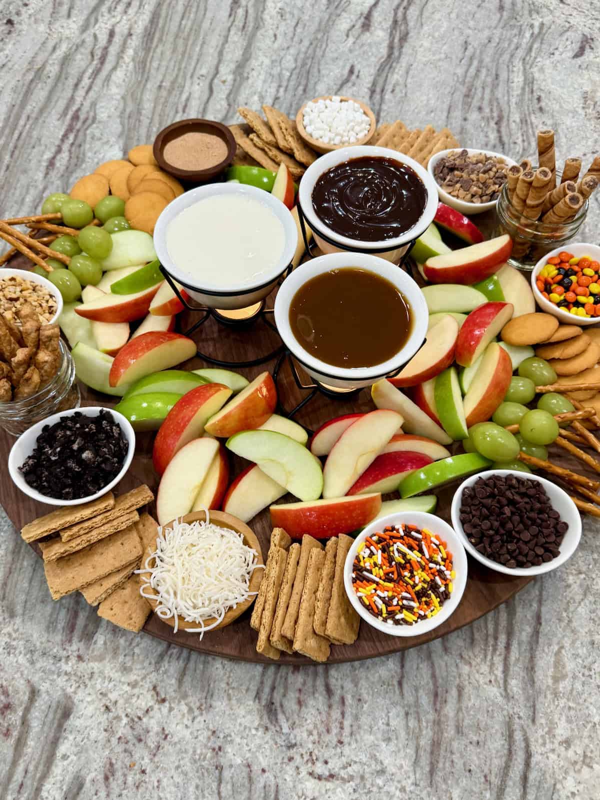 A large round food board filled with slices of red dandelion green apples and an assortment of dipping sauces and garnishes.