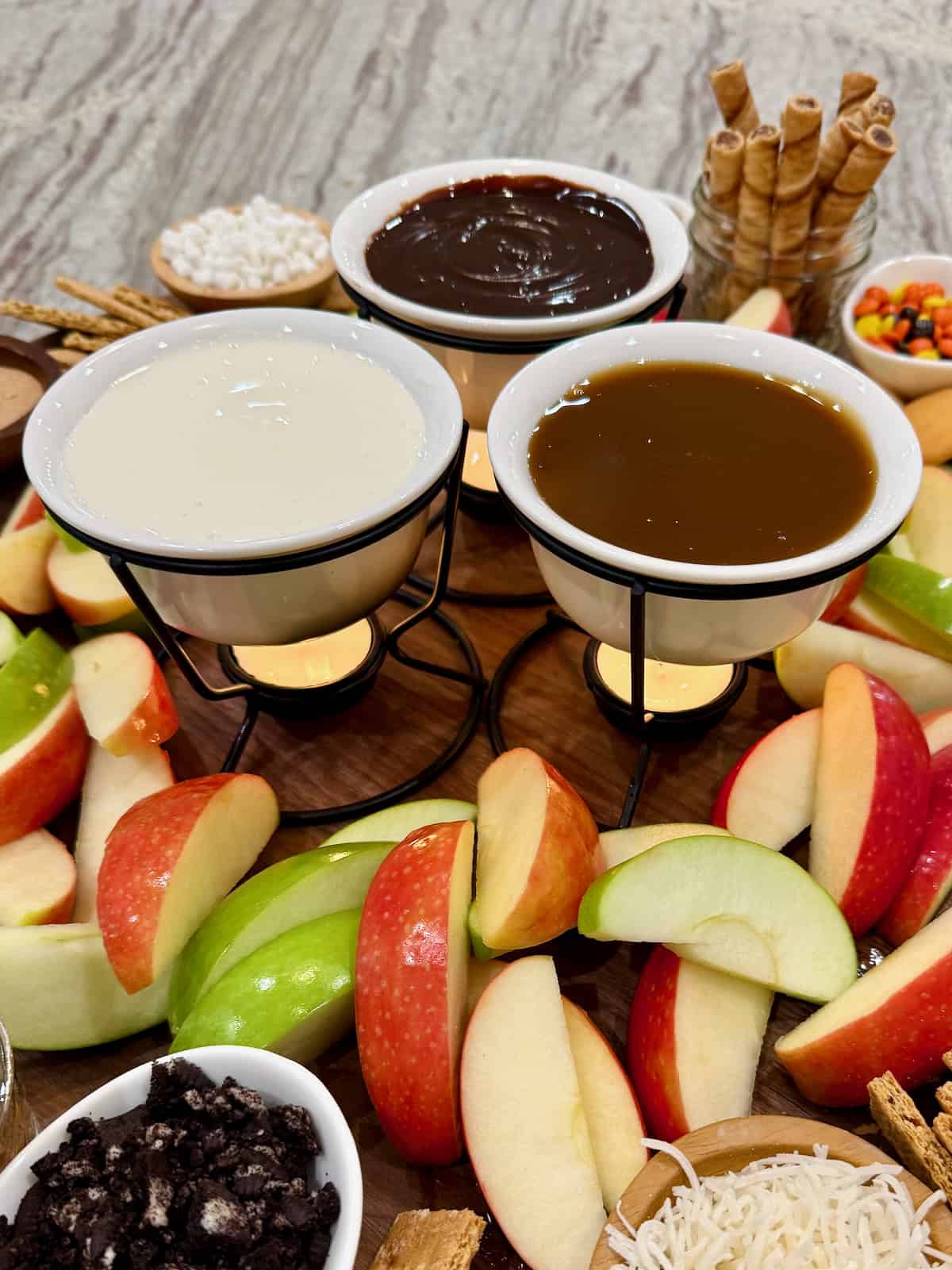 Fondue pots filled with hot fudge, marshmallow creme and caramel sauce on a food board surrounded by apple slices.