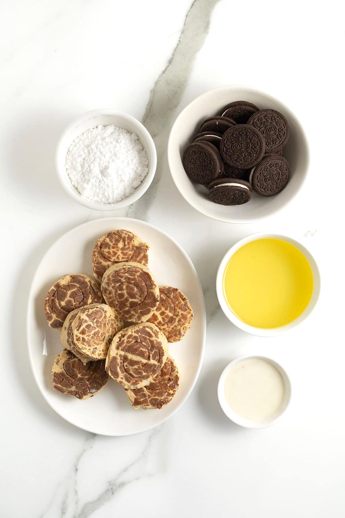 ingredients to make cinnamon roll deep fried Oreos in small white dishes on a white marble counter.