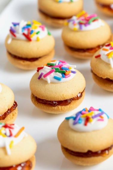 Eight vanilla wafer sandwich cookies topped with white icing and confetti sprinkles on a parchment surface.