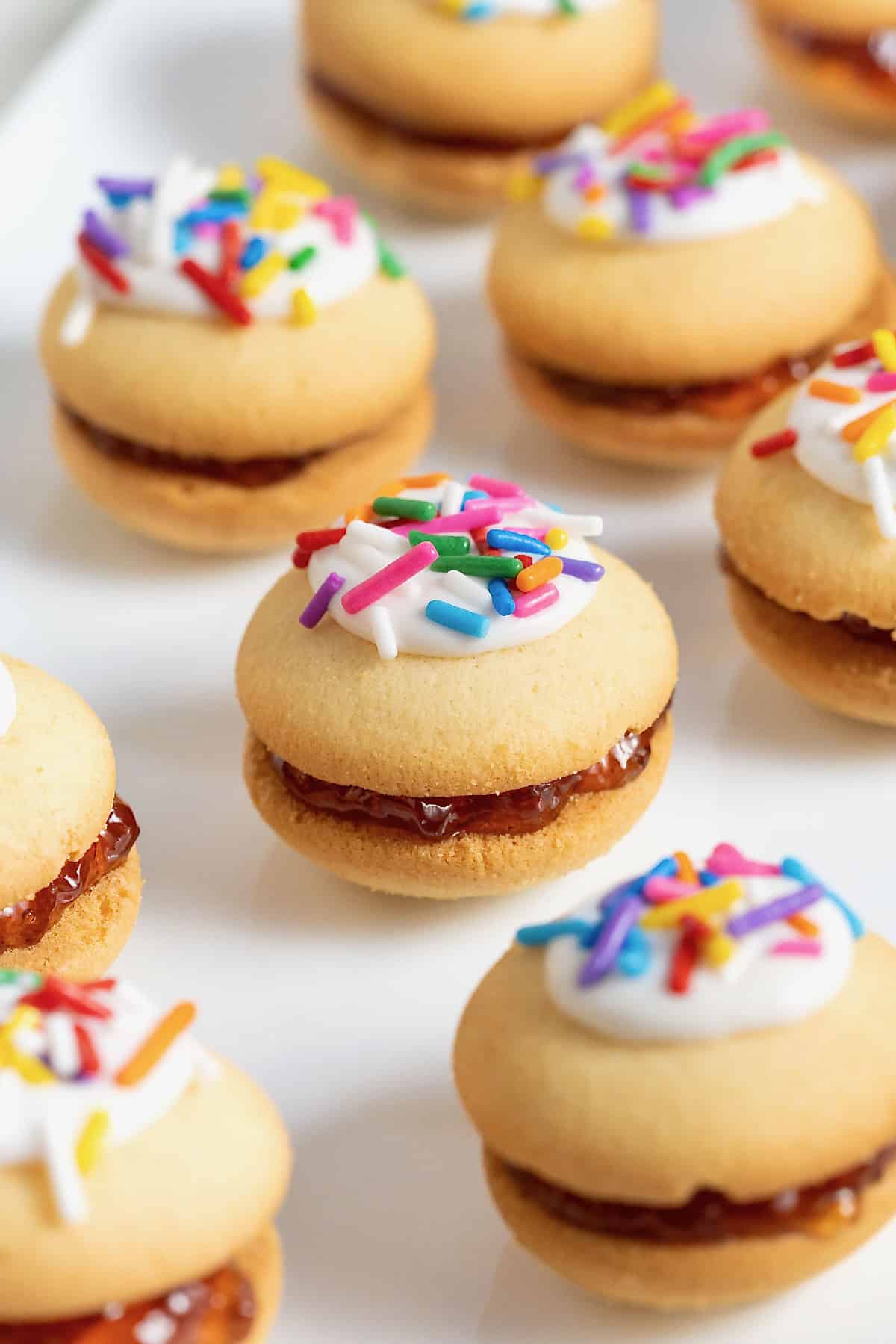 Eight vanilla wafer sandwich cookies topped with white icing and confetti sprinkles on a parchment surface.