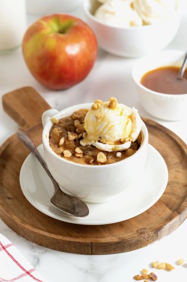 An apple cake baked in a white mug on a round wood cutting board. There is a scoop of ice cream on the cake.