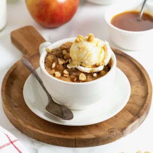 An apple cake in a white mug on a round wooden cutting board.