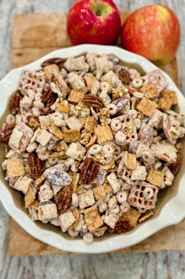 A white scalloped bowl filled with apple pie snack mix on a wooden cutting board next to two apples.