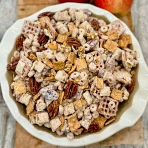A white scalloped bowl filled with apple pie snack mix on a wooden cutting board next to two apples.