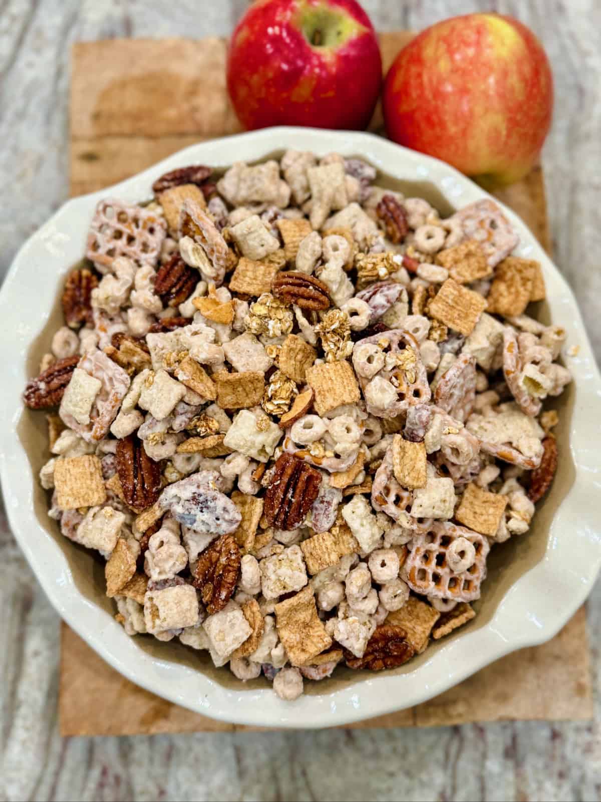 A white scalloped bowl filled with apple pie snack mix on a wooden cutting board next to two apples.
