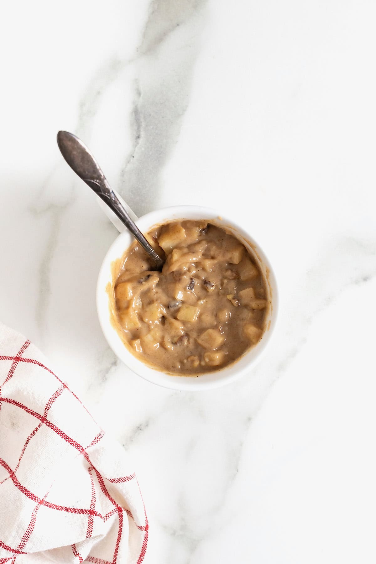 Apple cake batter with a spoon in it in a white mug on a white marble counter.