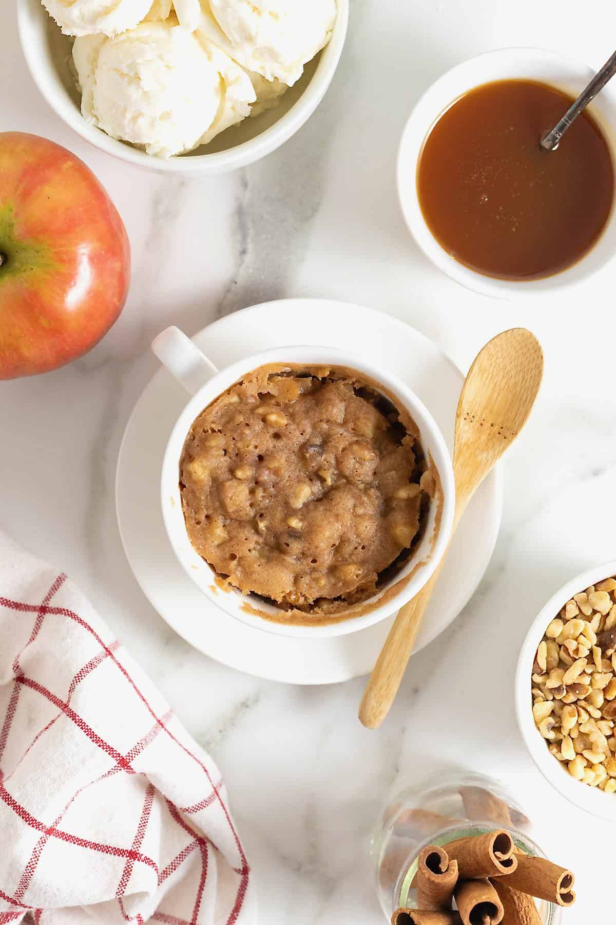 An apple cake in a white mug on a white plate with a wooden spoon next to it. It's surrounded by a small dish of vanilla ice cream, a small white dish of chopped walnuts and a small white dish of warm caramel sauce.