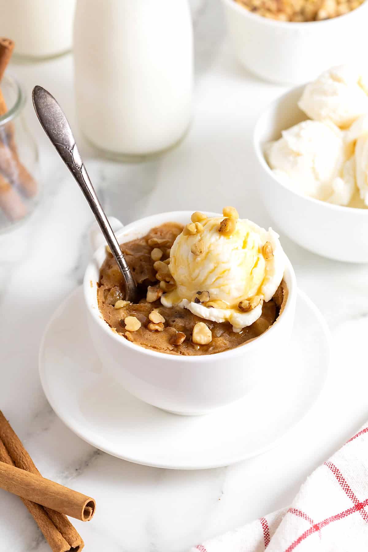 An apple cake baked in a white mug on a white plate. There is a scoop of ice cream on the cake and a spoon in the cake.
