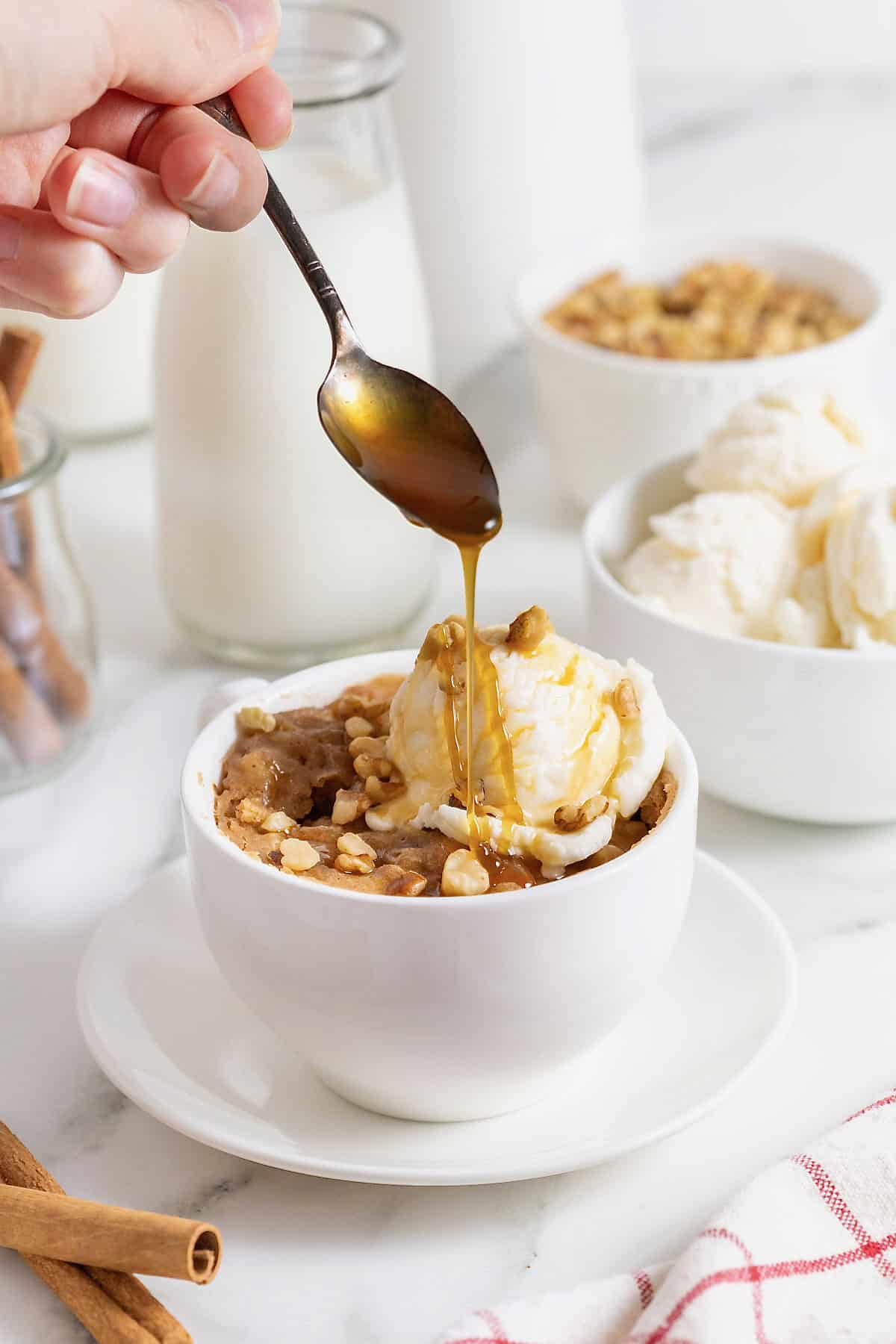 An apple cake baked in a white mug on a white plate. There is a scoop of ice cream on the cake and caramel being drizzled on the ice cream.