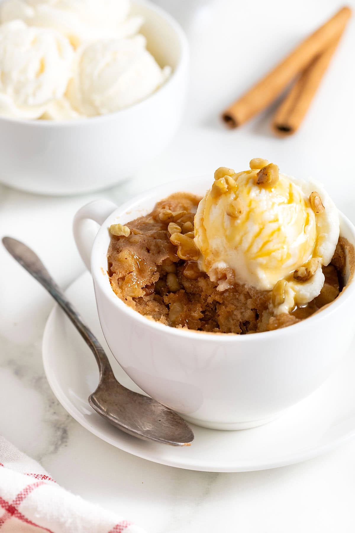 An apple cake baked in a white mug on a round wood cutting board. There is a scoop of ice cream on the cake. The cake has a bite taken out of it.