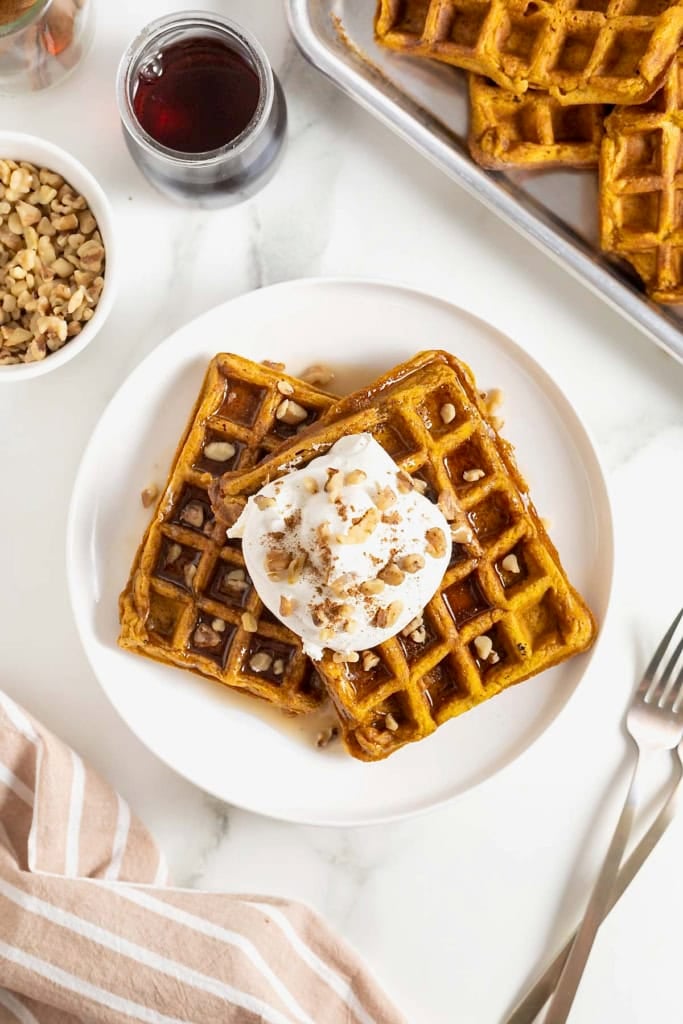 Two pumpkin waffles laying overlapping on a white plate. There is maple syrup on the waffles topped with whipped cream and chopped nuts.