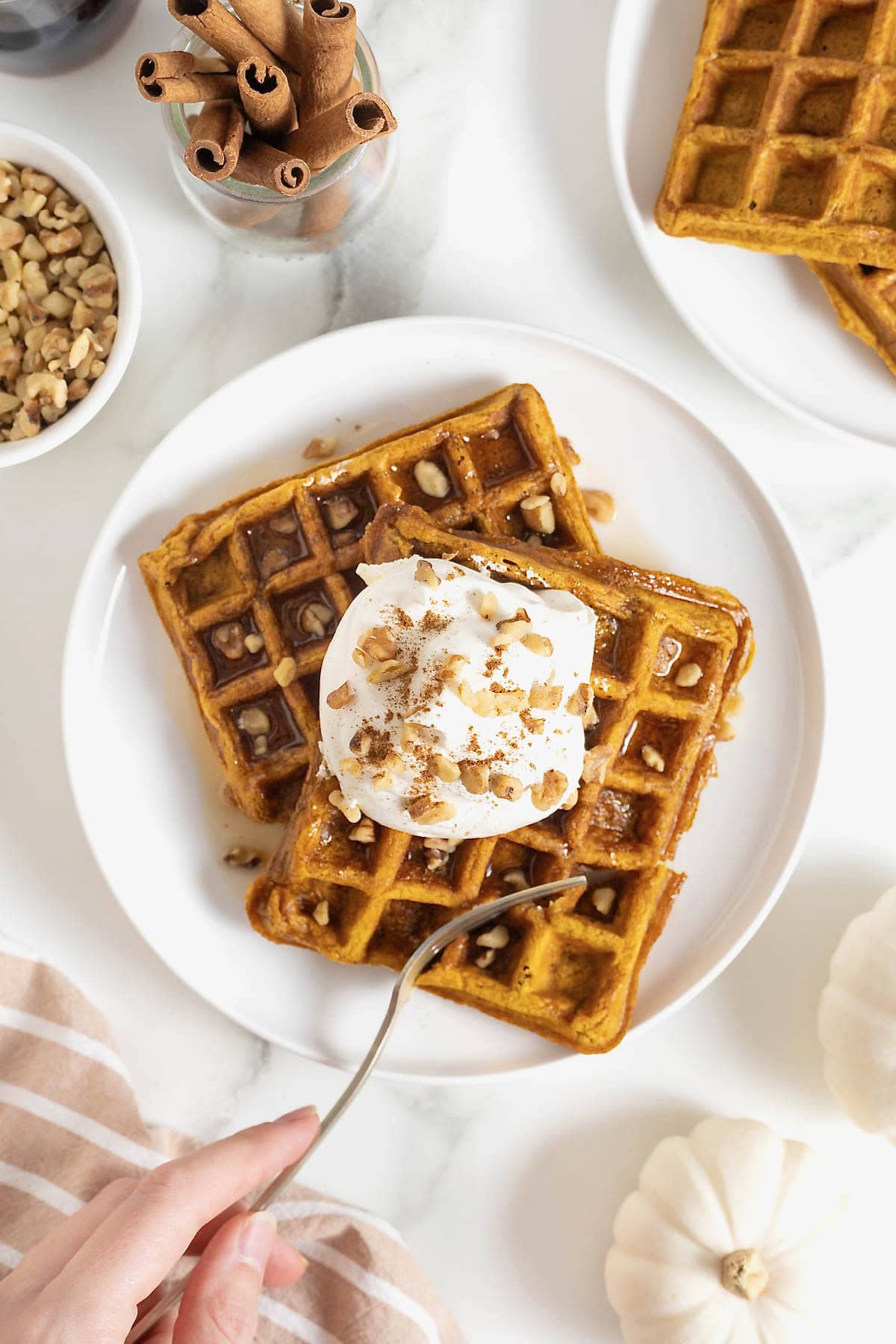 Two pumpkin waffles laying overlapping on a white plate. There is maple syrup on the waffles topped with whipped cream and chopped nuts.