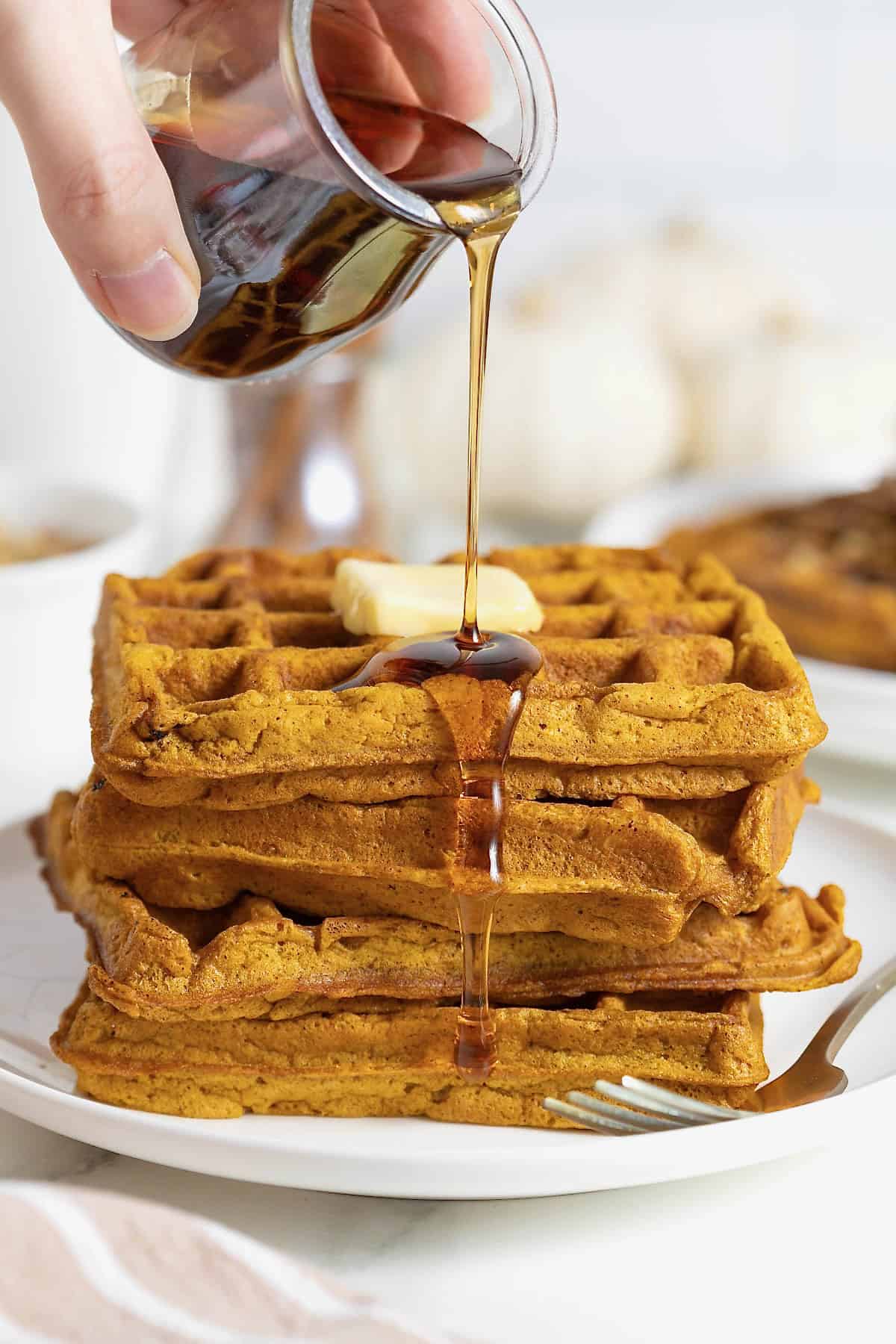 Maple syrup being poured onto a stack of five pancakes on a white plate.