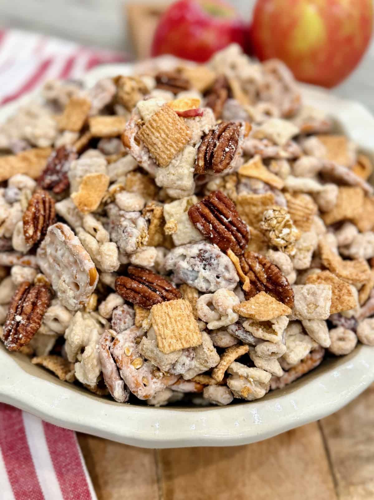 A white scalloped bowl filled with apple pie snack mix on a wooden cutting board next to two apples.