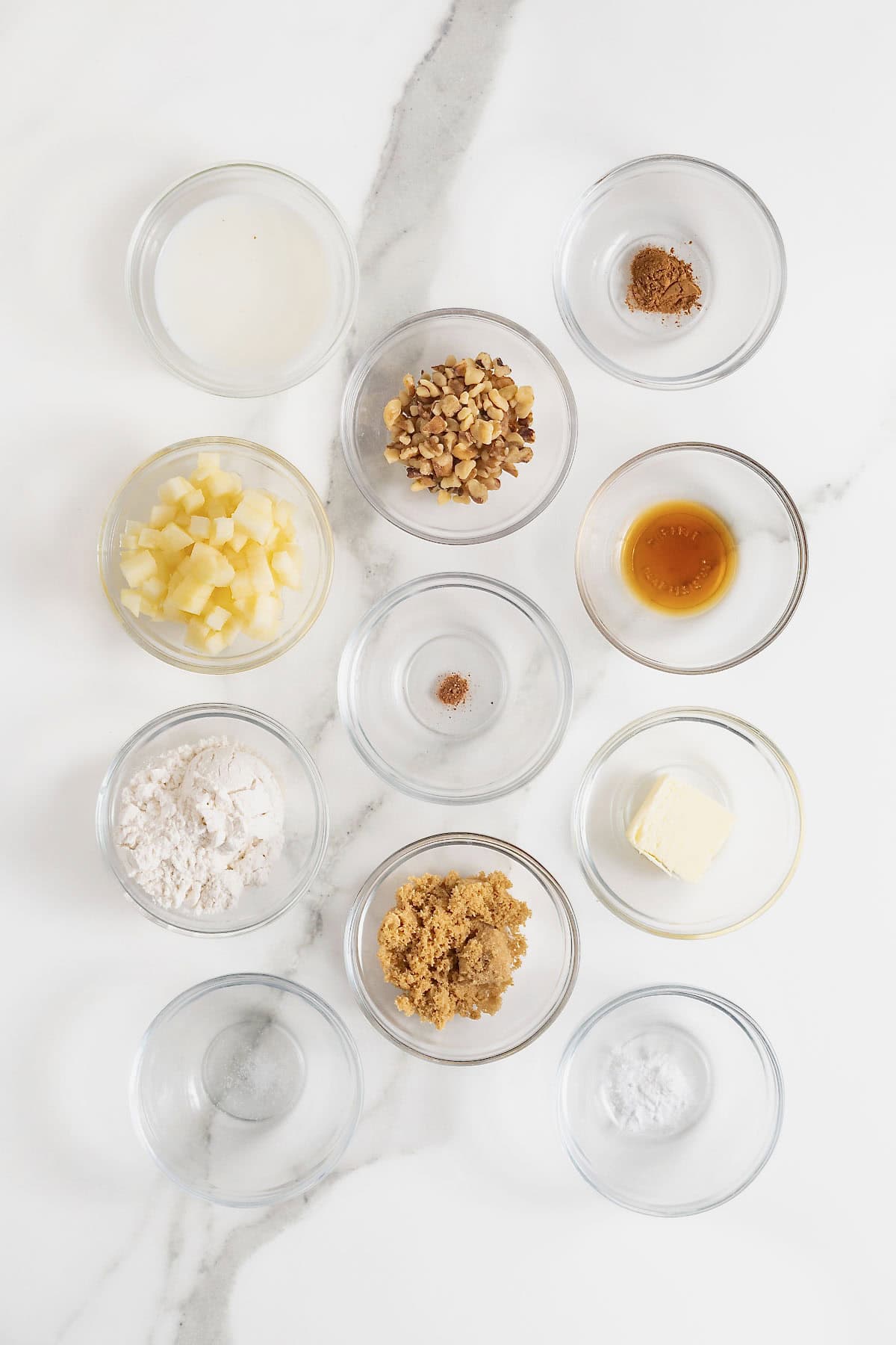 Ingredients to make a microwave apple cake in a mug in small glass dishes on a white marble counter.