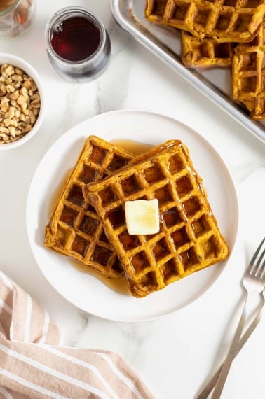 Two pumpkin waffles laying overlapping on a white plate. There is a square of butter on the top waffle.