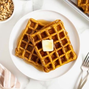 Two pumpkin waffles laying overlapping on a white plate. There is a square of butter on the top waffle.