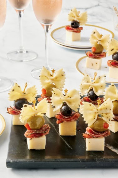 7 appetizer bites made with cheese cubes topped with salami slices, black and green olives and a piece of bowtie pasta arranged on a black serving tray.