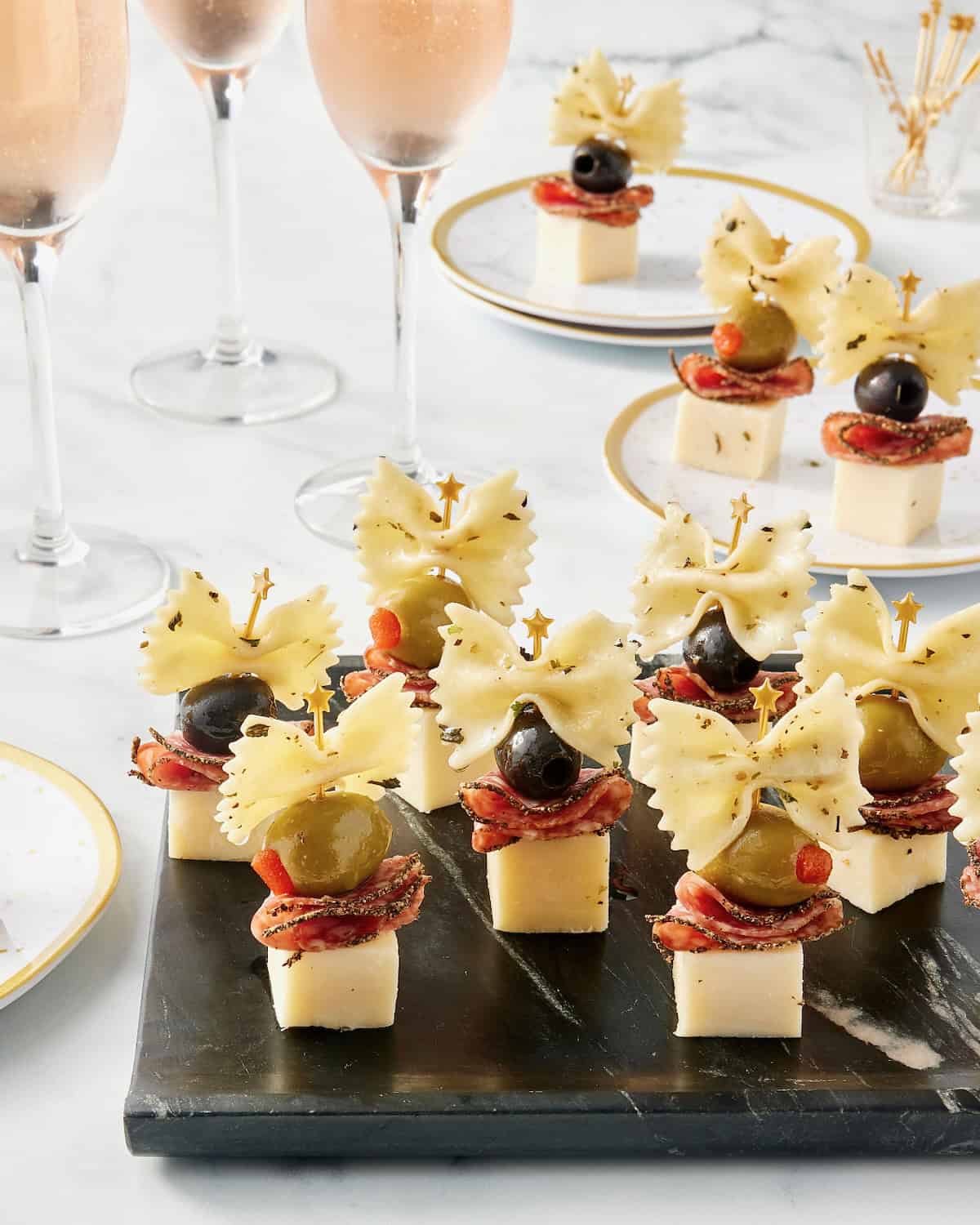 7 appetizer bites made with cheese cubes topped with salami slices, black and green olives and a piece of bowtie pasta arranged on a black serving tray.