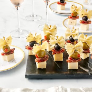 7 appetizer bites made with cheese cubes topped with salami slices, black and green olives and a piece of bowtie pasta arranged on a black serving tray.