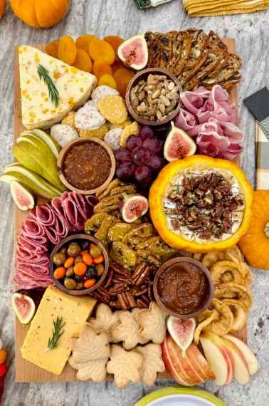 A rectangular wooden food board filled with autumnal snacks like cheese, salami, sliced pear and a pumpkin dip.