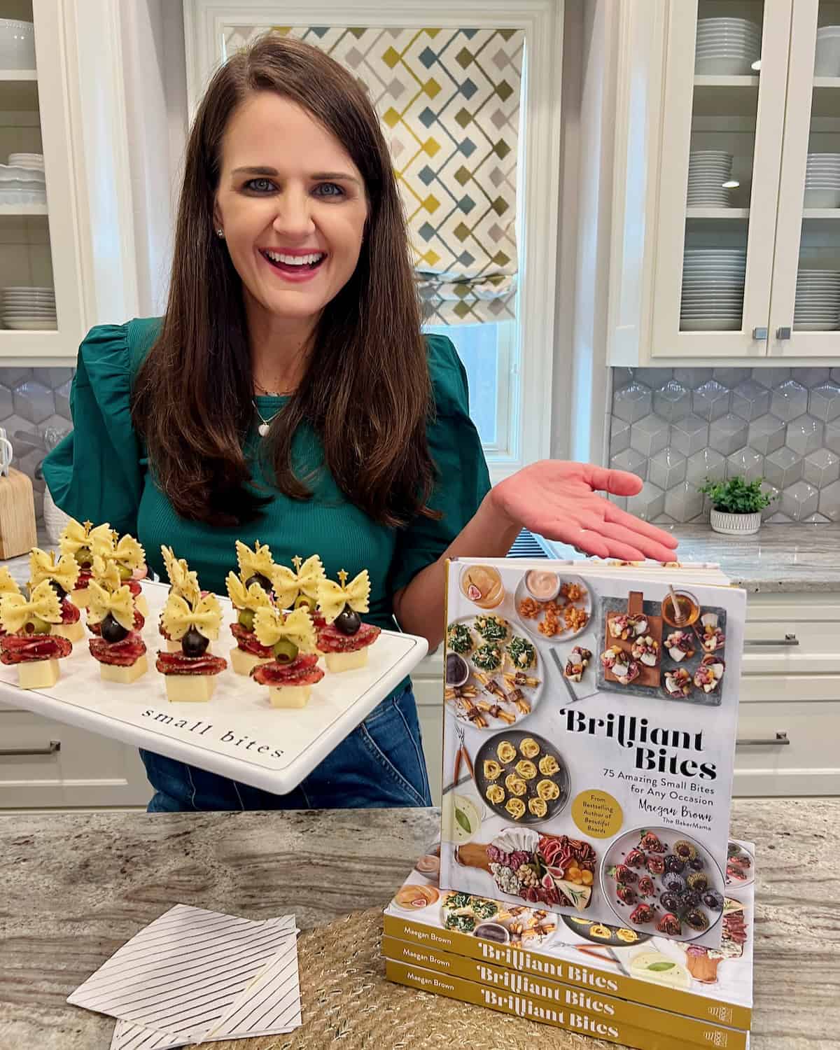 Cookbook author Maegan Brown holding a plate of bow-tie antipasto bites and showing off her cookbook, Brilliant Bites.