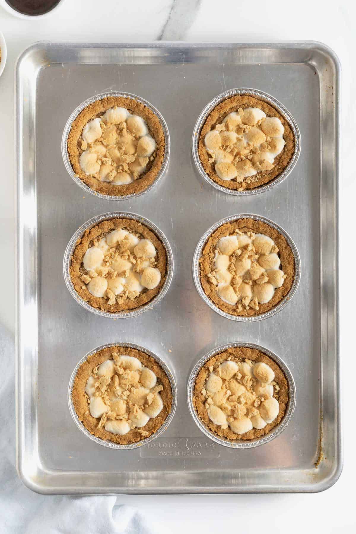 Six s'mores cookie pies topped with graham cracker crumbs on an aluminum baking sheet.