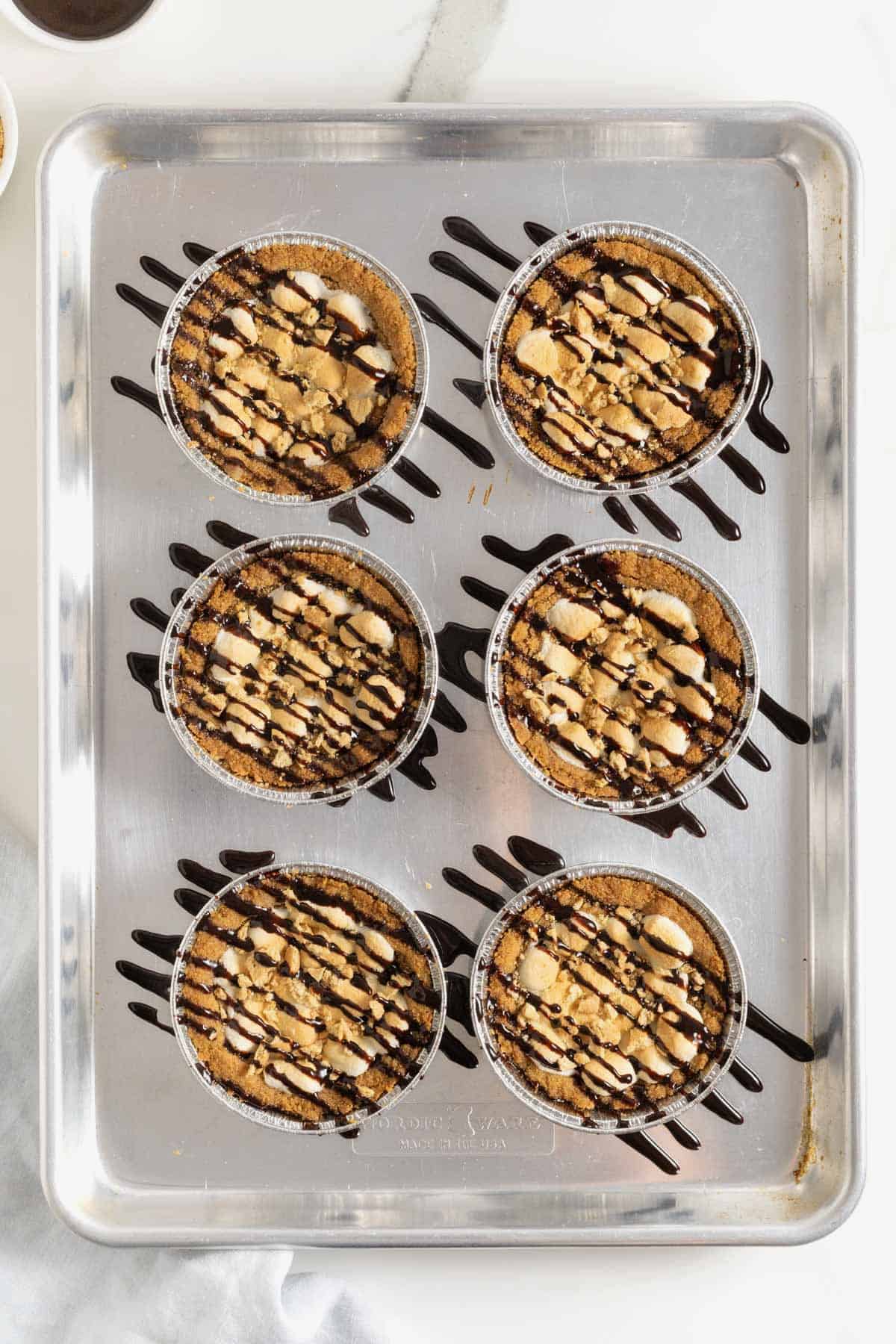 Six s'mores cookie pies drizzled with chocolate sauce on an aluminum baking sheet.
