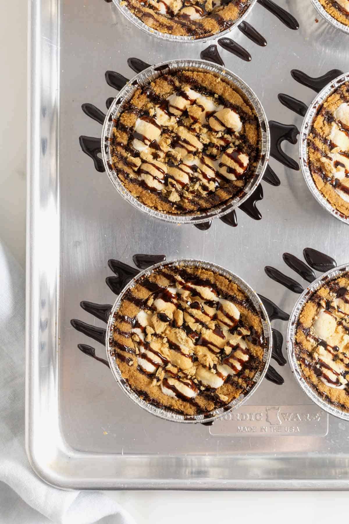 Five s'mores cookie pies drizzled with chocolate sauce on an aluminum baking sheet.
