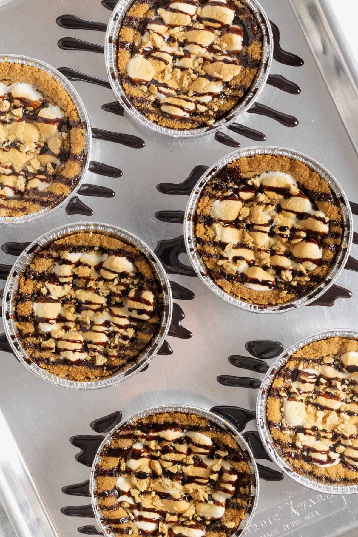 Six s'mores cookie pies drizzled with chocolate sauce on an aluminum baking sheet.