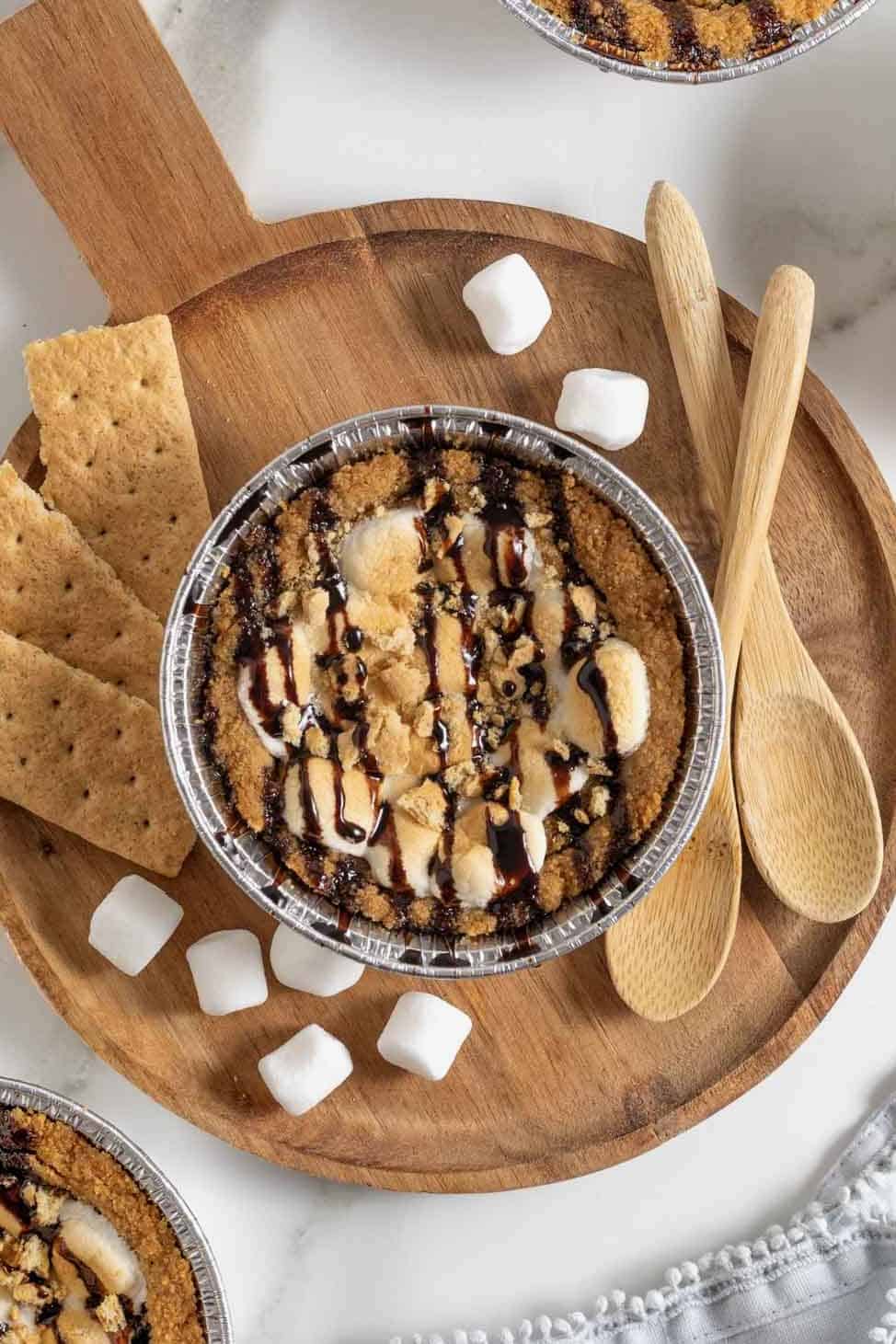 A mini s'mores cookie pie on a round wood cutting board surrounded by mini marshmallows, graham crackers and two wooden spoons.