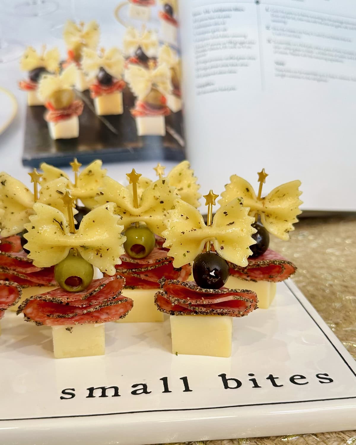 Appetizer bites made with cheese cubes topped with salami slices, black and green olives and a piece of bowtie pasta arranged on a white ceramic serving tray with the words "small bites" on it.