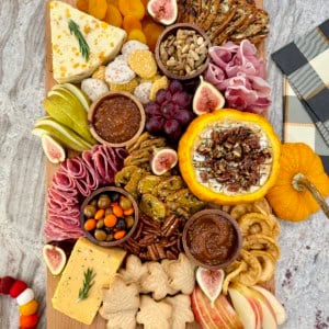 A rectangular wooden food board filled with autumnal snacks like cheese, salami, sliced pear and a pumpkin dip.