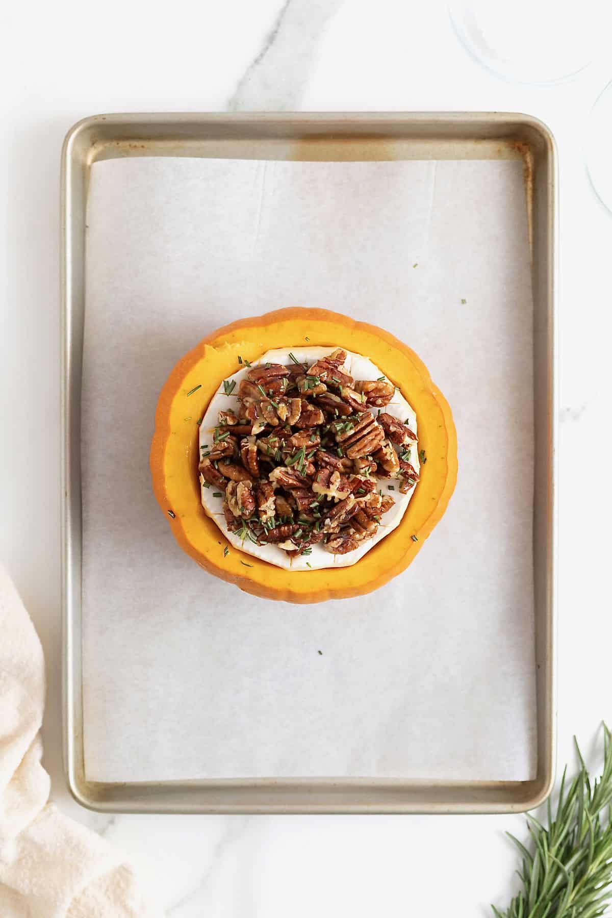 Brie in a mini pumpkin topped with maple pecans and rosemary on a parchment lined baking sheet.