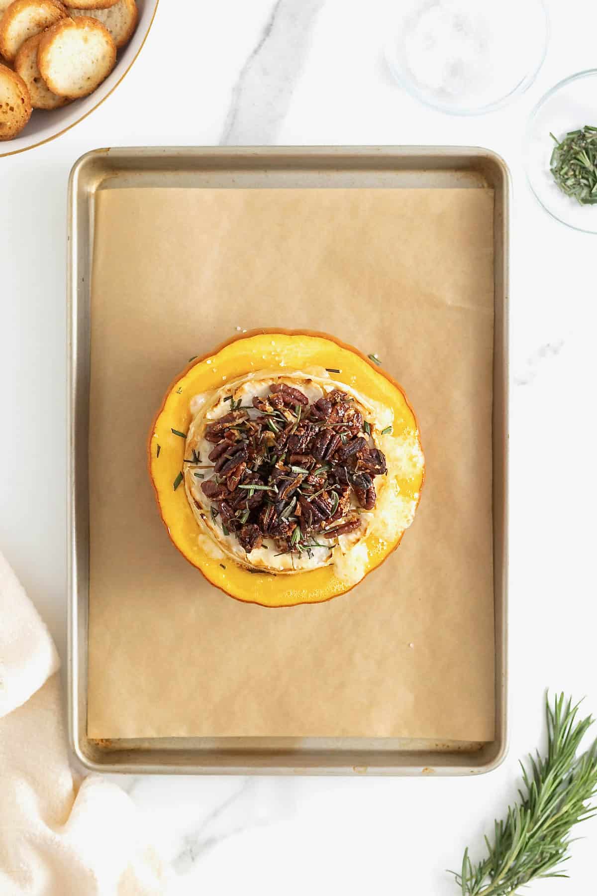 Baked brie in a mini pumpkin topped with maple pecans and rosemary on a parchment lined baking sheet.