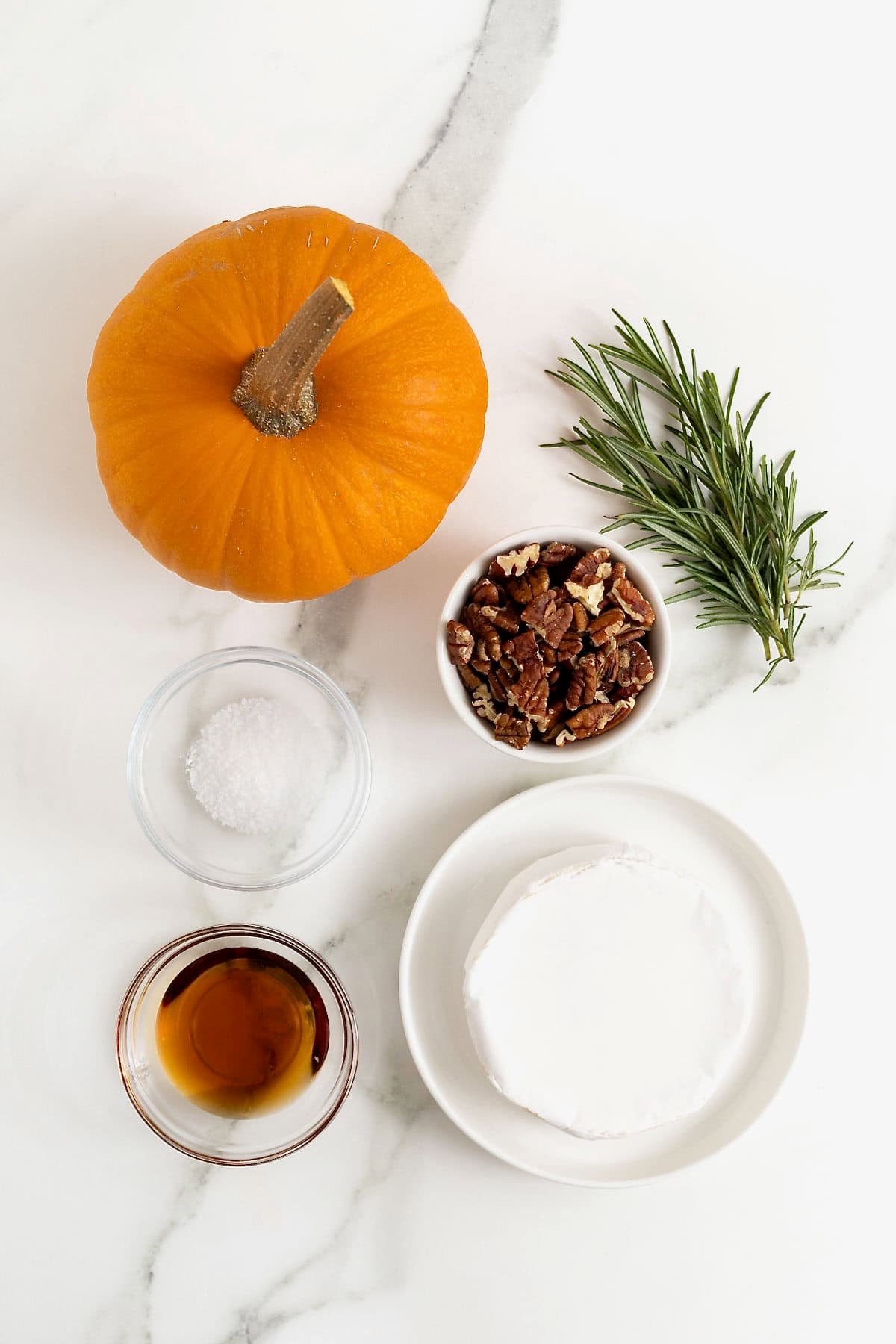 Ingredients to make pumpkin baked brie with maple, pecans and rosemary on white marble counter.
