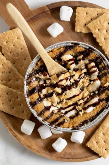 A mini s'mores cookie pie on a wooden cutting board surrounded by mini marshmallows and graham cracker pieces.