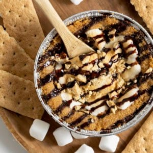 A mini s'mores cookie pie on a wooden cutting board surrounded by mini marshmallows and graham cracker pieces.