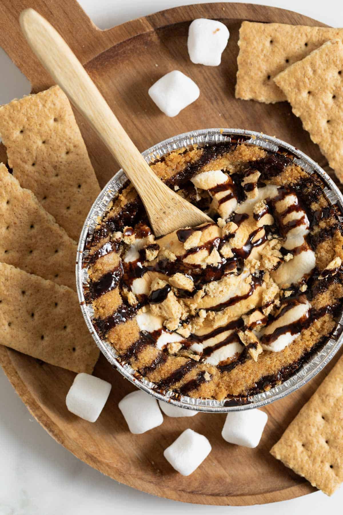 A mini s'mores cookie pie on a wooden cutting board surrounded by mini marshmallows and graham cracker pieces.